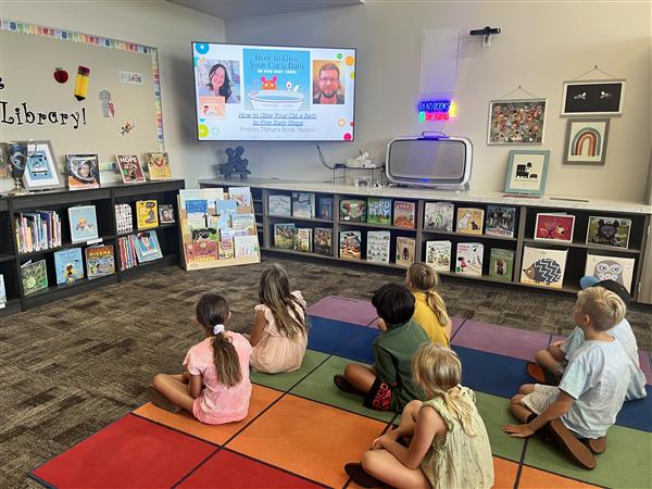 A small group of students sits on a rug, looking at TV Screen where teacher has projected a book title