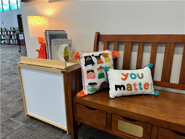 A small bench in the library with a pillow that says You Matter next to books and a lamp